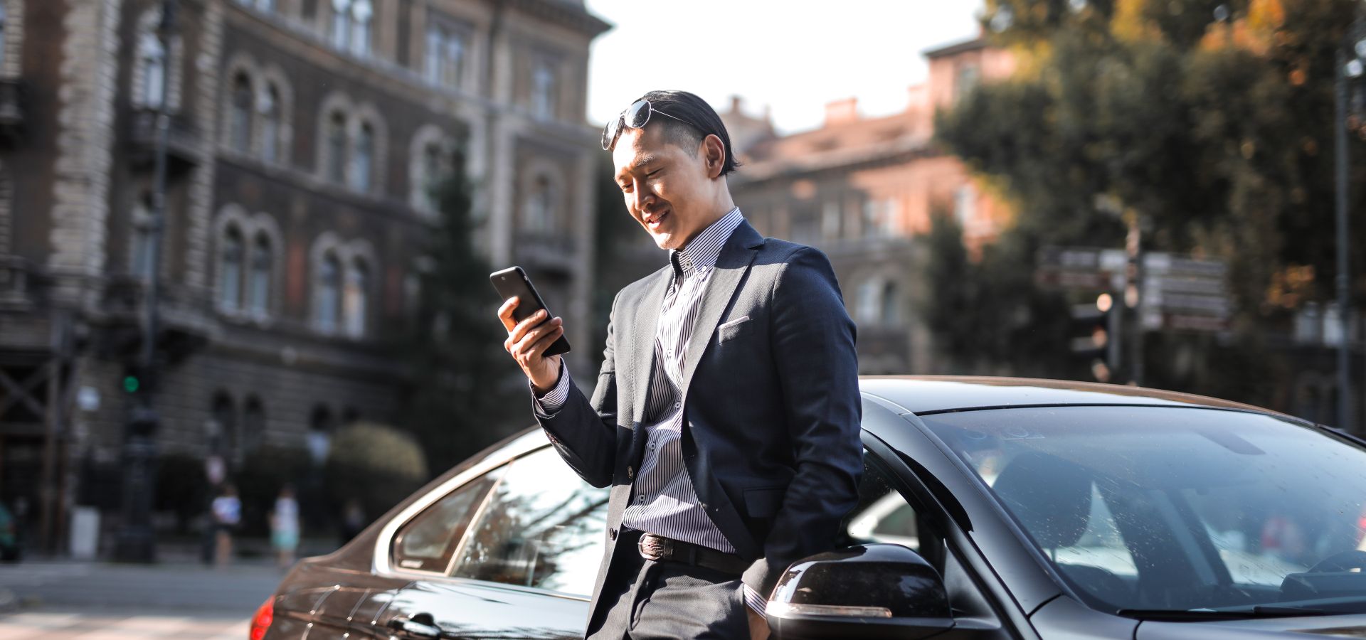 Smiling sales professional in a suit checking his phone beside his car